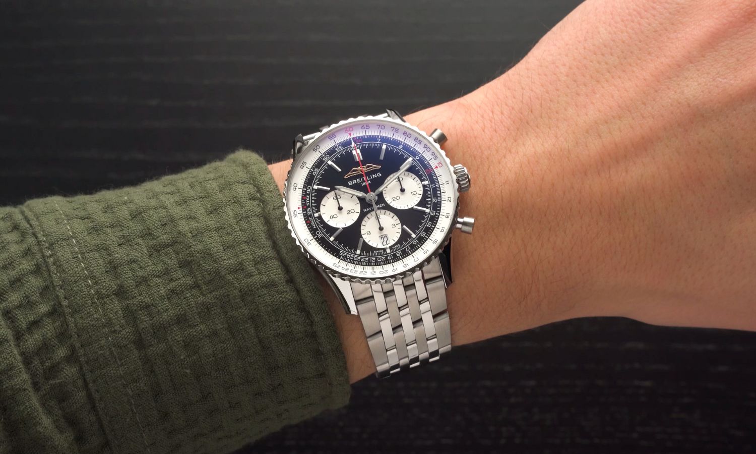 Breitling Navitimer B01 Chronograph 43 mit schwarzem Zifferblatt am Edelstahlarmband an einem Handgelenk mit dunkelgrünem Ärmel getragen vor schwarzem Hintergrund