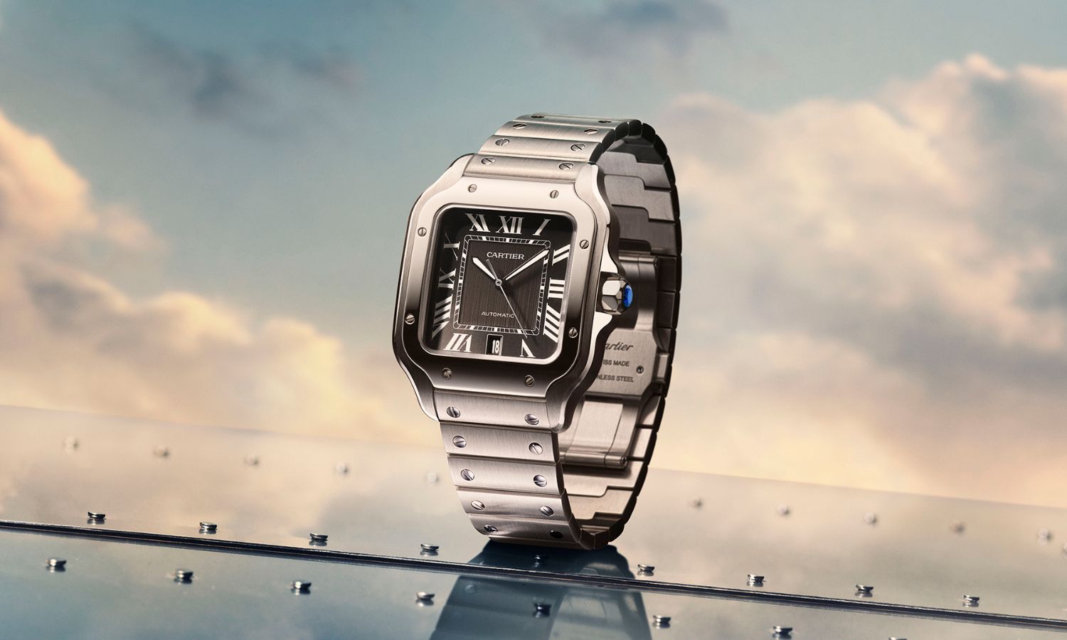 Cartier Santos Referenz WSSA0096 mit schwarzem Zifferblatt am Edelstahlarmband stehend auf einem Metallboden vor blauem Himmel mit Wolken