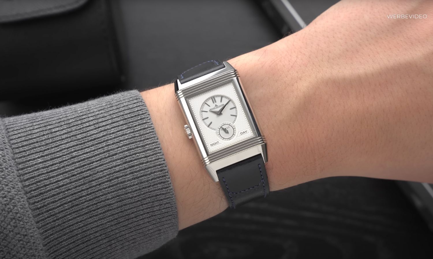 Jaeger-LeCoultre Reverso mit weißem Zifferblatt am schwarzen Lederarmband an einem Handgelenk mit grauem Ärmel getragen vor schwarzem Hintergrund