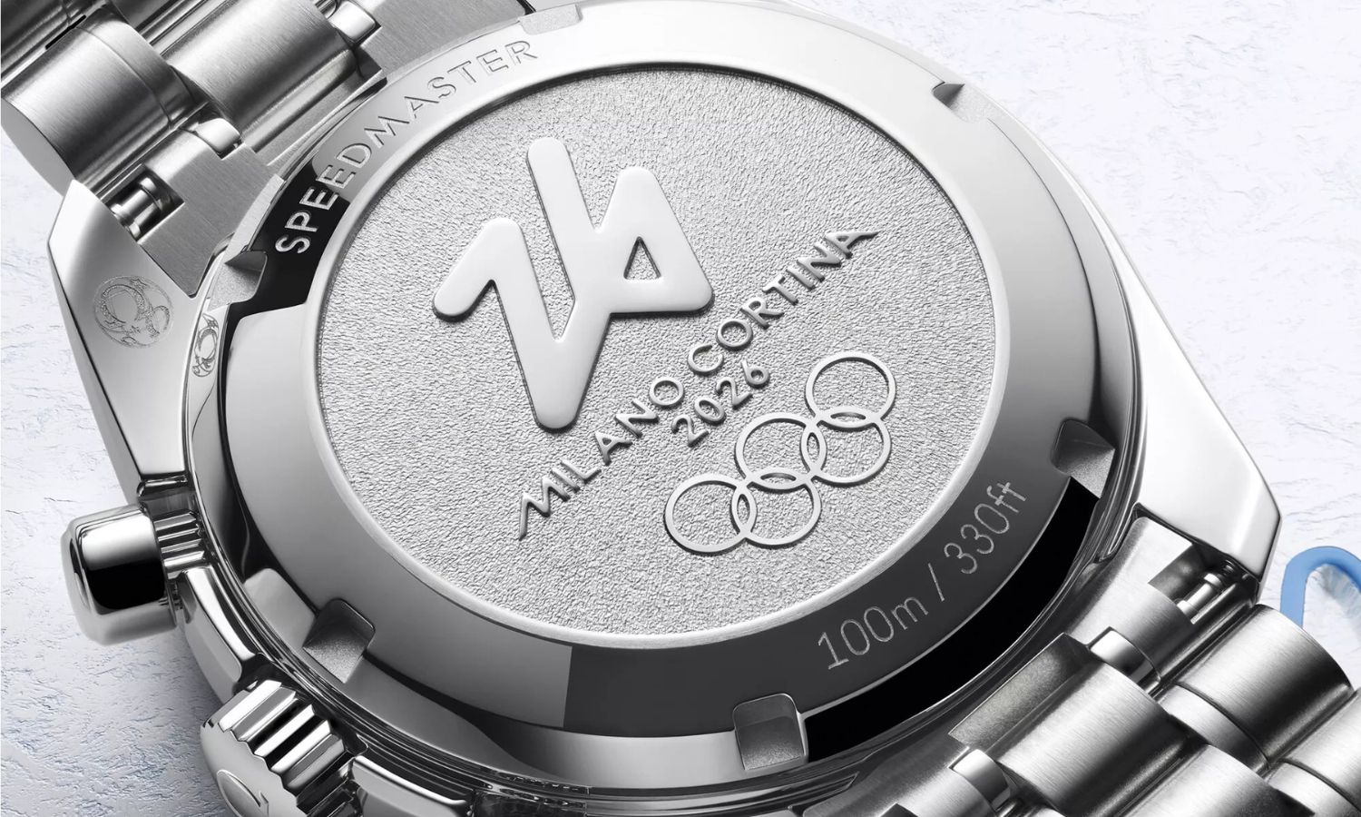 Der Gehäuseboden der OMEGA Speedmaster Milano Cortina 2026 Referenz 522.30.38.50.04.001 am Edelstahlarmband von hinten abgebildet vor weißem Hintergrund mit blauen Verzierungen