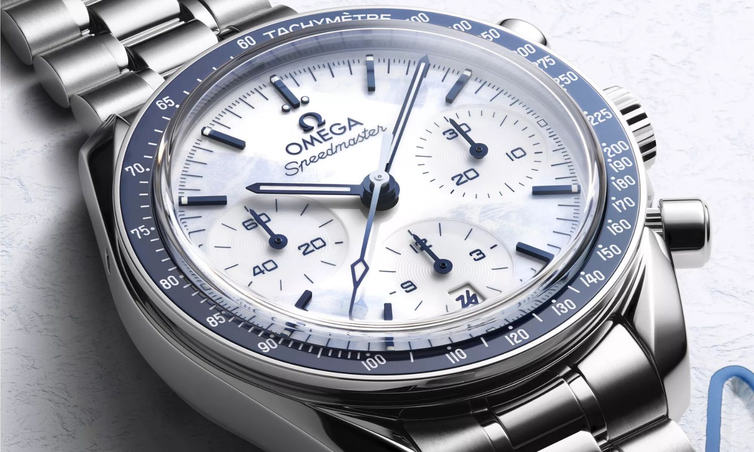 Das weiße Zifferblatt der OMEGA Speedmaster Milano Cortina 2026 Referenz 522.30.38.50.04.001 am Edelstahlarmband von vorne abgebildet vor weißem Hintergrund mit blauen Verzierungen