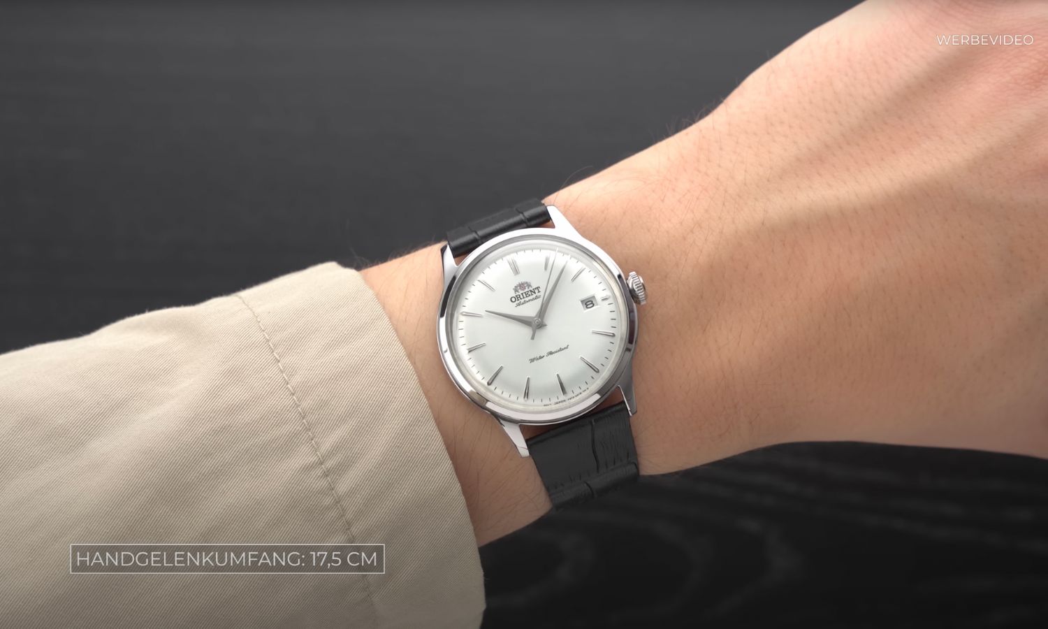 Orient Bambino mit weißem Zifferblatt am schwarzen Lederarmband an einem Handgelenk mit beigem Ärmel getragen vor schwarzem Hintergrund