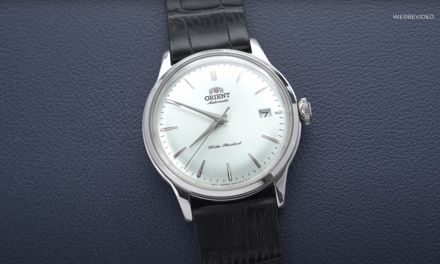 Orient Bambino mit weißem Zifferblatt am schwarzen Lederarmband liegend vor dunkelblauem Hintergrund