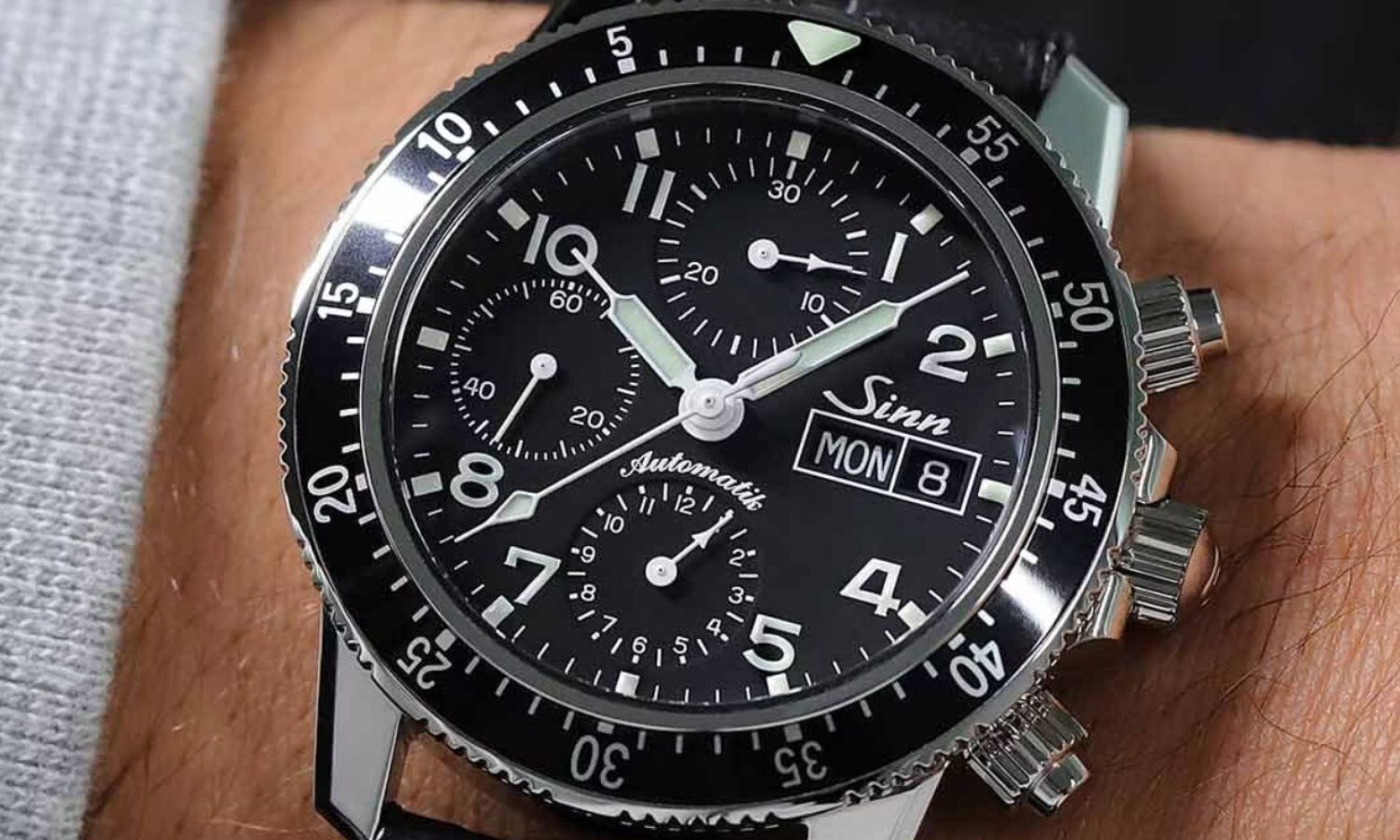 Sinn 103 St Sa mit schwarzem Zifferblatt am braunen Lederarmband an einem Handgelenk mit hellgrauem Ärmel getragen