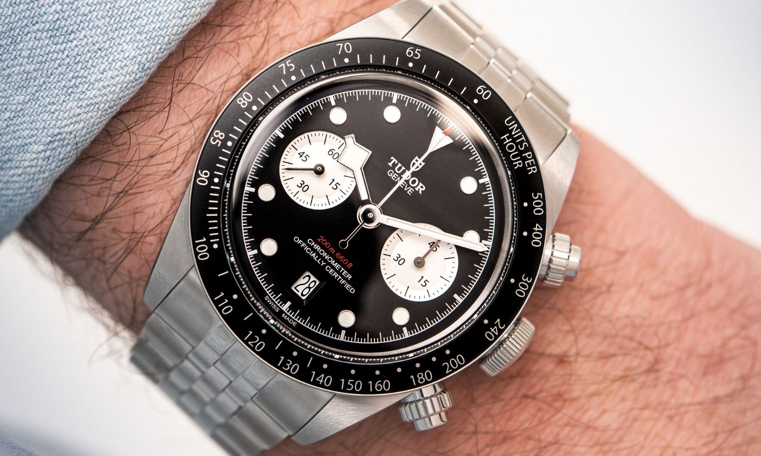 TUDOR Black Bay Chrono mit schwarzem Zifferblatt am Edelstahlarmband an einem Handgelenk mit hellblauem Ärmel getragen vor weißem Hintergrund