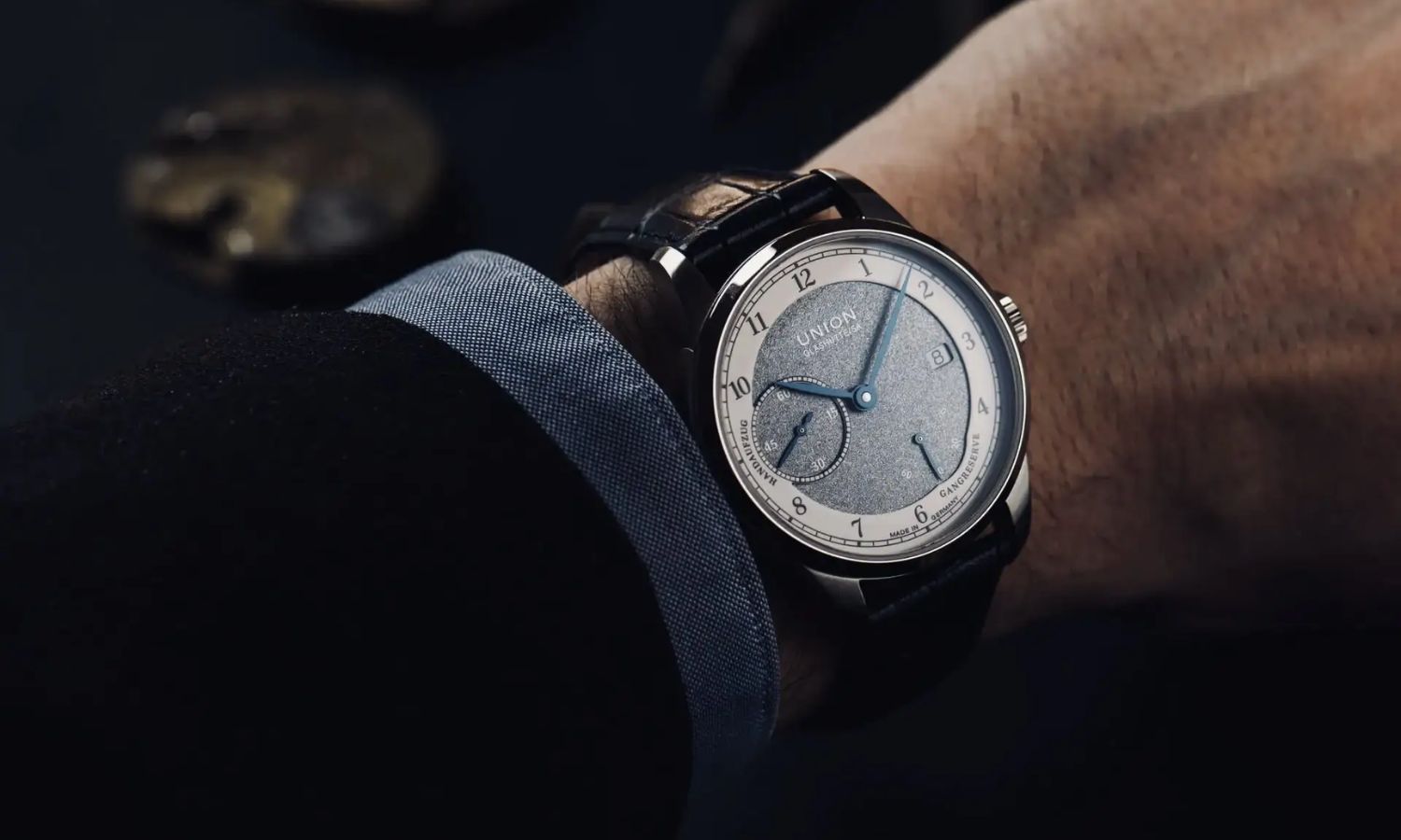 Union Glashütte 1893 Johannes Dürrstein Edition Gangreserve mit blaugrünem Zifferblatt am blauen Lederarmband an einem Handgelenk mit dunkelblauem Ärmel getragen vor dunklem Hintergrund
