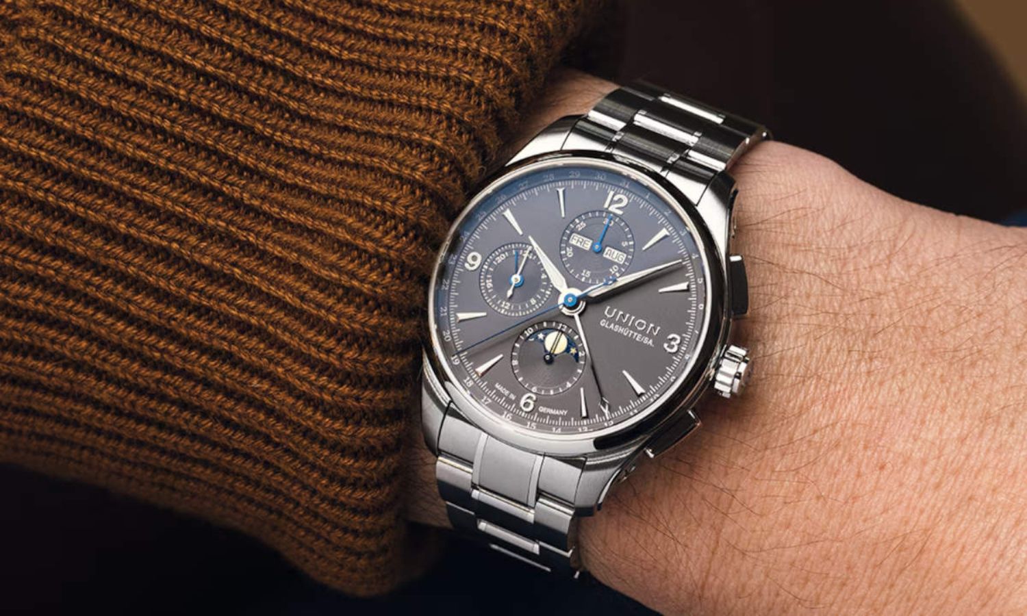 Union Glashütte Belisar Chronograph Moon Phase mit grauem Zifferblatt am Edelstahlarmband an einem Handgelenk mit braunem Ärmel getragen vor schwarzem Hintergrund