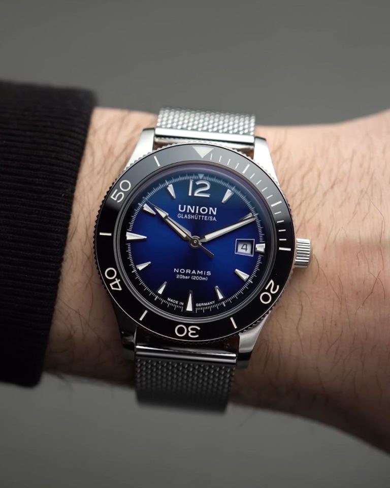 UNION Glashütte Noramis Datum Sport mit blauem Zifferblatt am Edelstahlarmband an einem Handgelenk mit schwarzem Ärmel getragen vor grauem Hintergrund