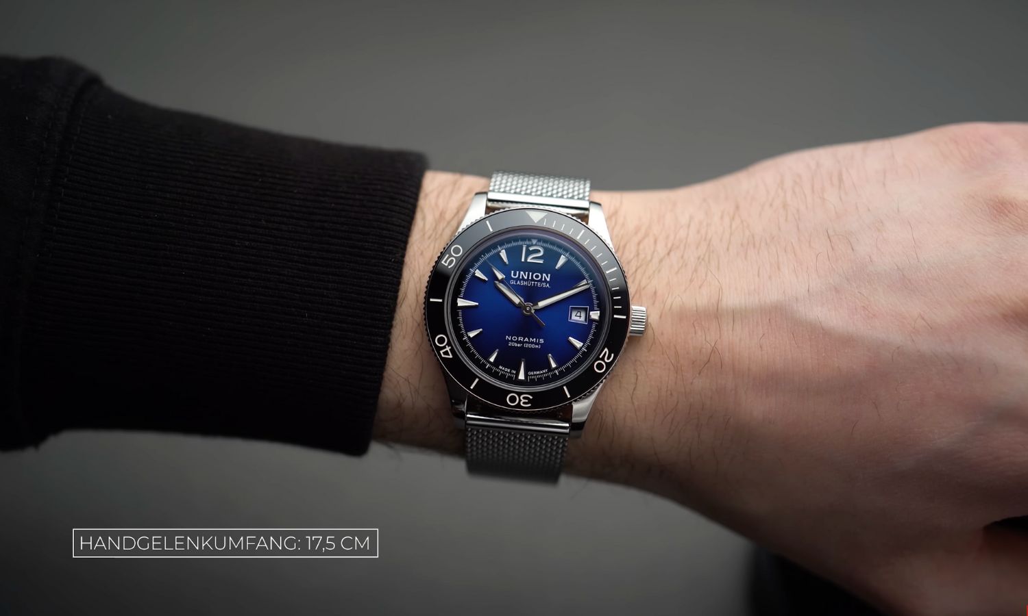UNION Glashütte Noramis Datum Sport mit blauem Zifferblatt am Edelstahlarmband an einem Handgelenk mit schwarzem Ärmel getragen vor grauem Hintergrund