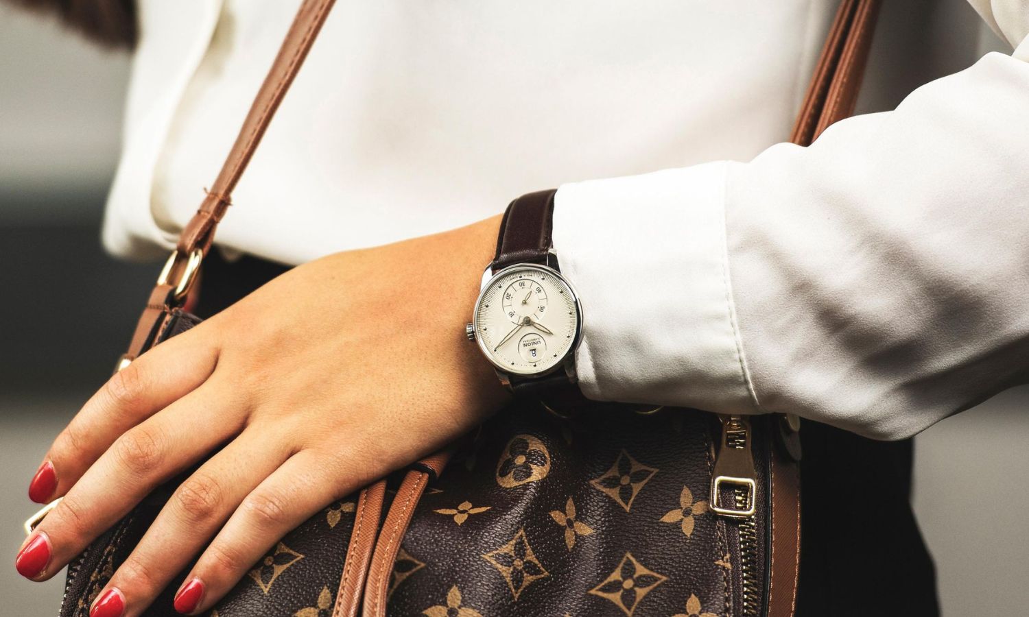 Union Glashütte Seris Kleine Sekunde mit silbernem Zifferblatt am braunen Lederarmband an einem Damenhandgelenk mit weißem Ärmel getragen vor einer braunen Louis Vuitton Tasche