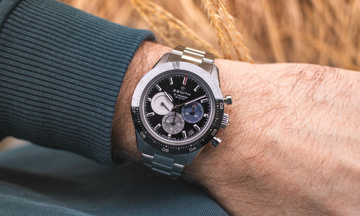 Zenith Chronomaster Sport mit schwarzem Zifferblatt am Edelstahlarmband an einem Handgelenk mit dunkelgrünem Ärmel getragen vor hellbraunem Hintergrund