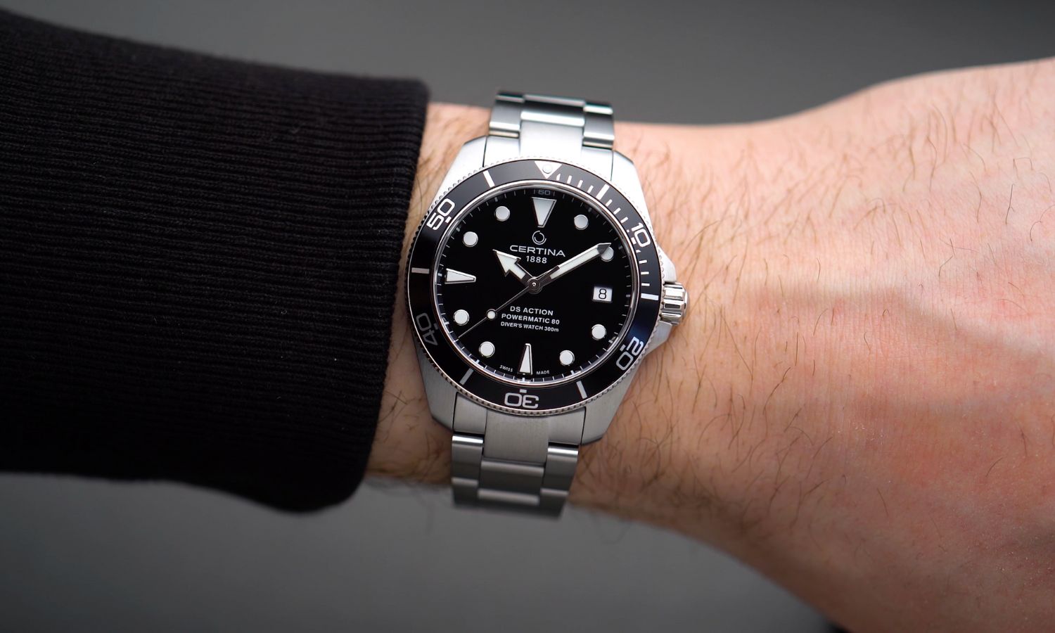 Certina DS Action Diver 38mm mit schwarzem Zifferblatt am Edelstahlarmband an einem Handgelenk mit schwarzem Ärmel getragen vor grauem Hintergrund