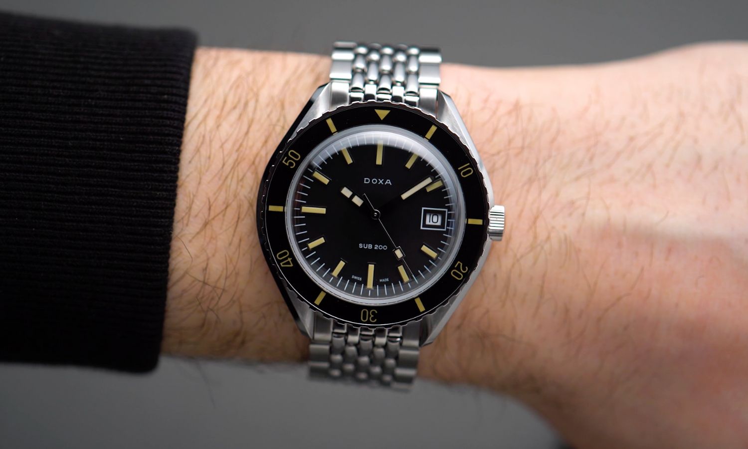 Doxa Sub 200T Sharkhunter mit schwarzem Zifferblatt am Edelstahlarmband an einem Handgelenk mit schwarzem Ärmel getragen vor grauem Hintergrund