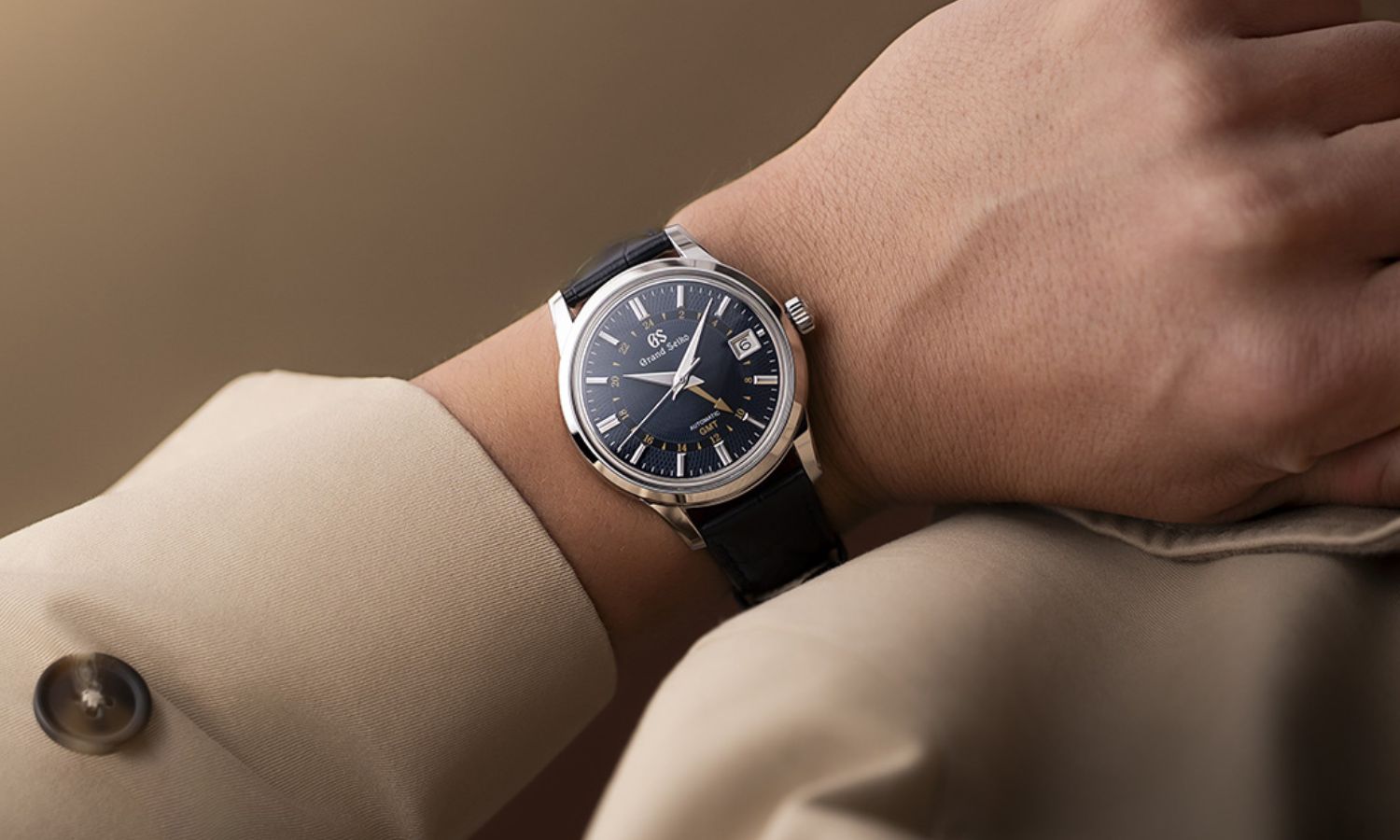 Grand Seiko SBGM257 „Moondrop“ mit dunkelblauem Zifferblatt am dunkelblauen Lederarmband an einem Handgelenk mit beigem Ärmel getragen vor hellbraunem Hintergrund