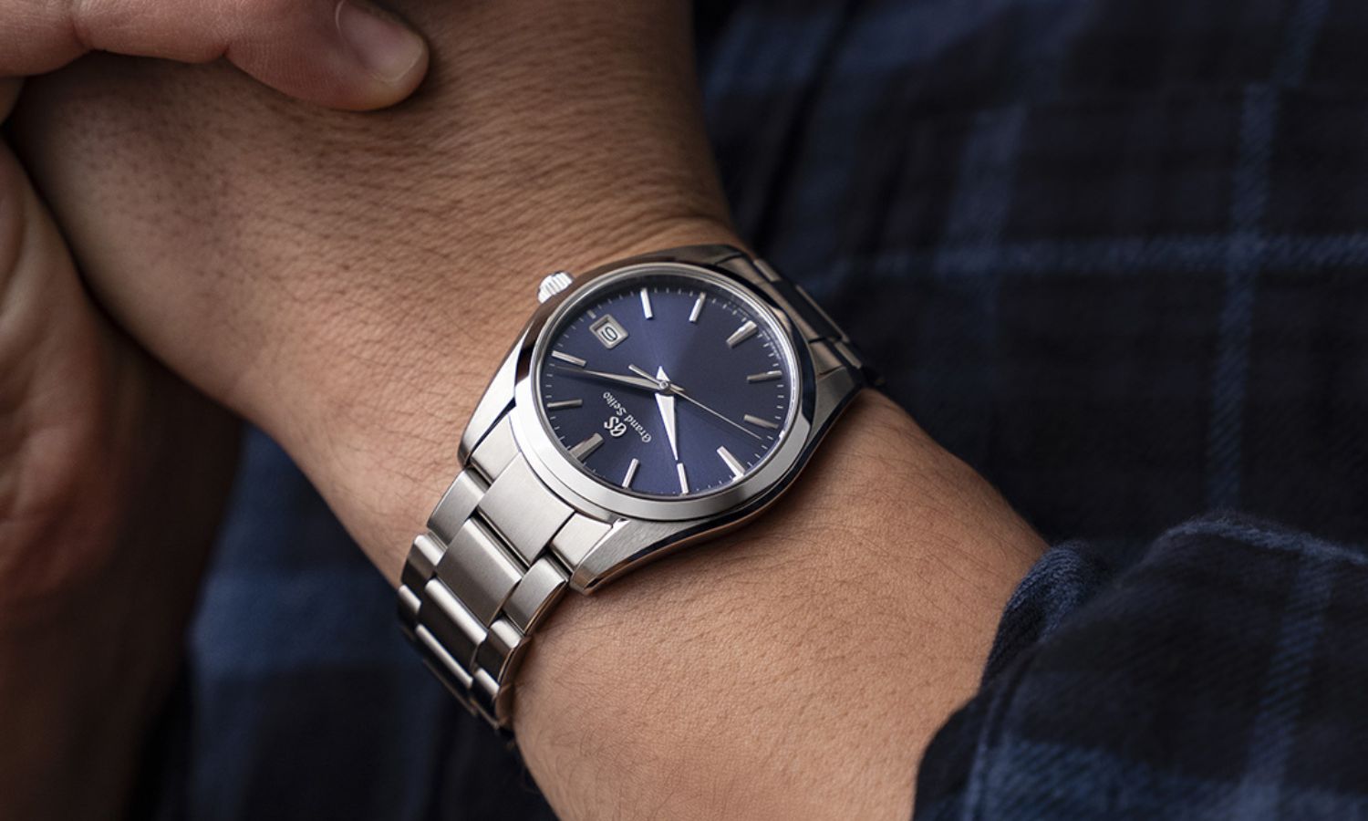 Grand Seiko SBGX265 mit dunkelblauem Zifferblatt am Edelstahlarmband an einem Handgelenk mit dunkelblauem Ärmel getragen vor dunklem Hintergrund