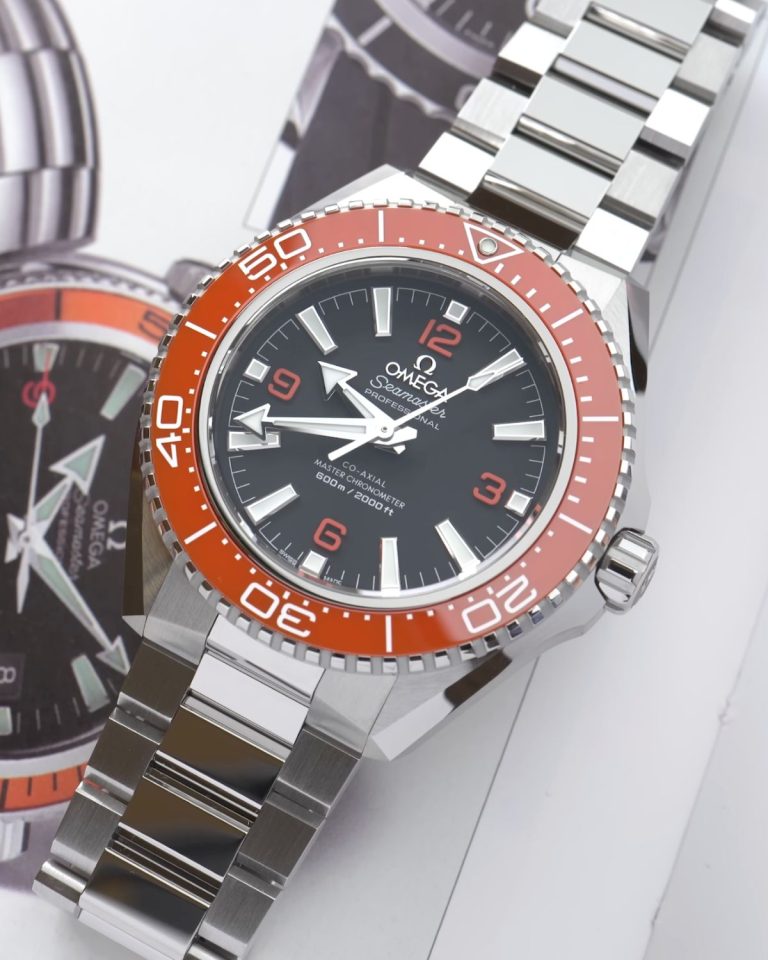 OMEGA Seamaster Planet Ocean 2025 mit schwarzem Zifferblatt und orangener Lünette am Edelstahlarmband liegend auf einem Magazin mit einem Bild älterer OMEGA Seamaster Planet Ocean Modelle