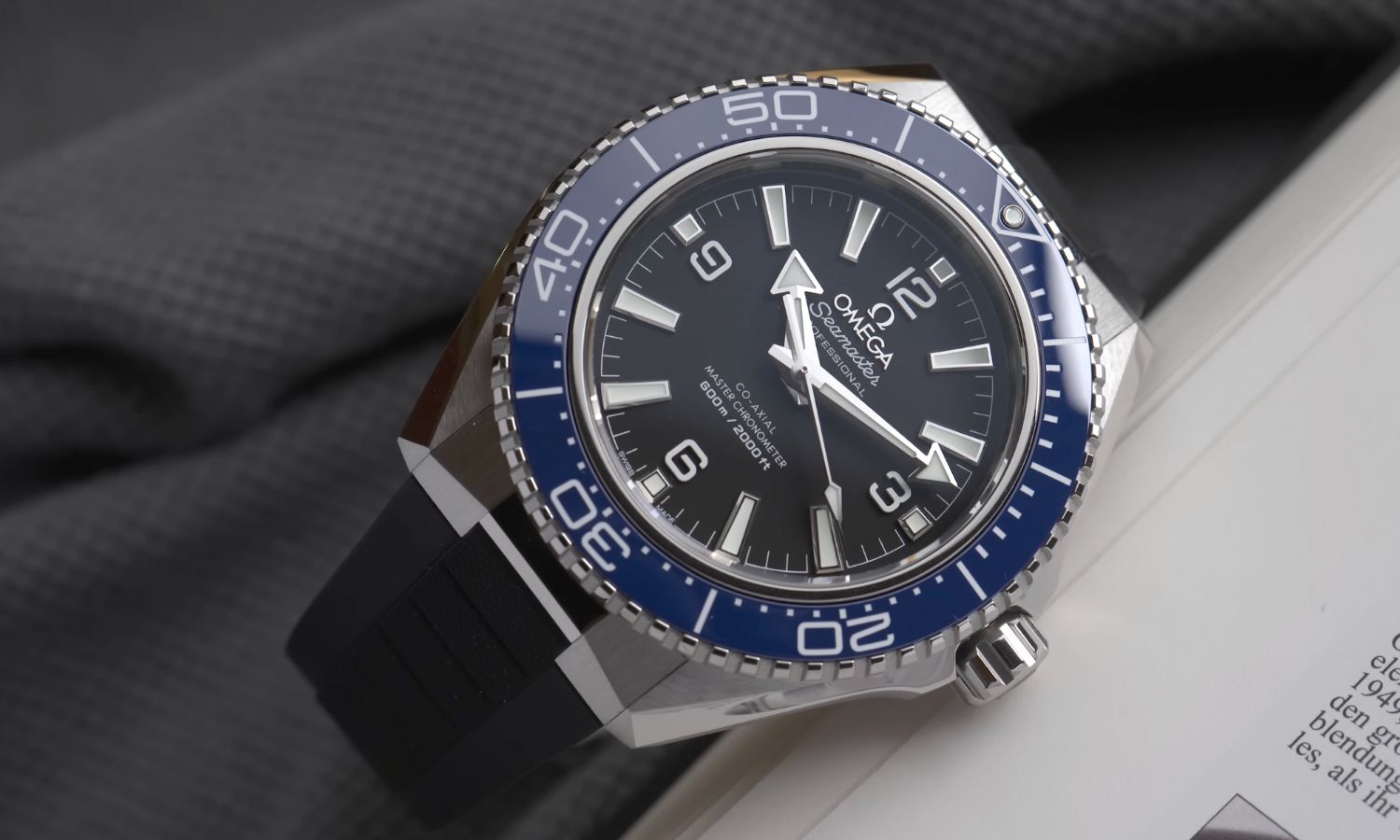 OMEGA Seamaster Planet Ocean 2025 mit schwarzem Zifferblatt und blauer Lünette am schwarzen Kautschukarmband seitlich liegend an ein Buch gelehnt vor grauem Hintergrund