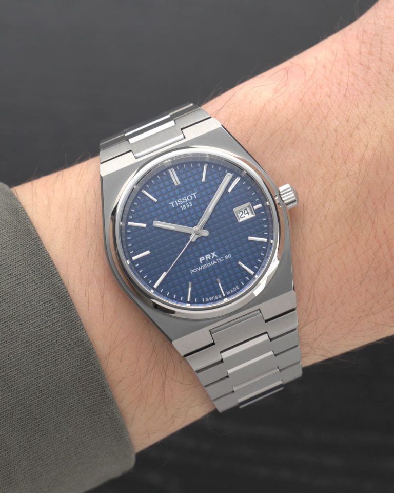 Tissot PRX Powermatic 80 Titan 38 mm Referenz T137.807.44.041.00 mit blauem Zifferblatt am Titanarmband an einem Handgelenk mit olivgrünem Ärmel getragen vor schwarzem Hintergrund