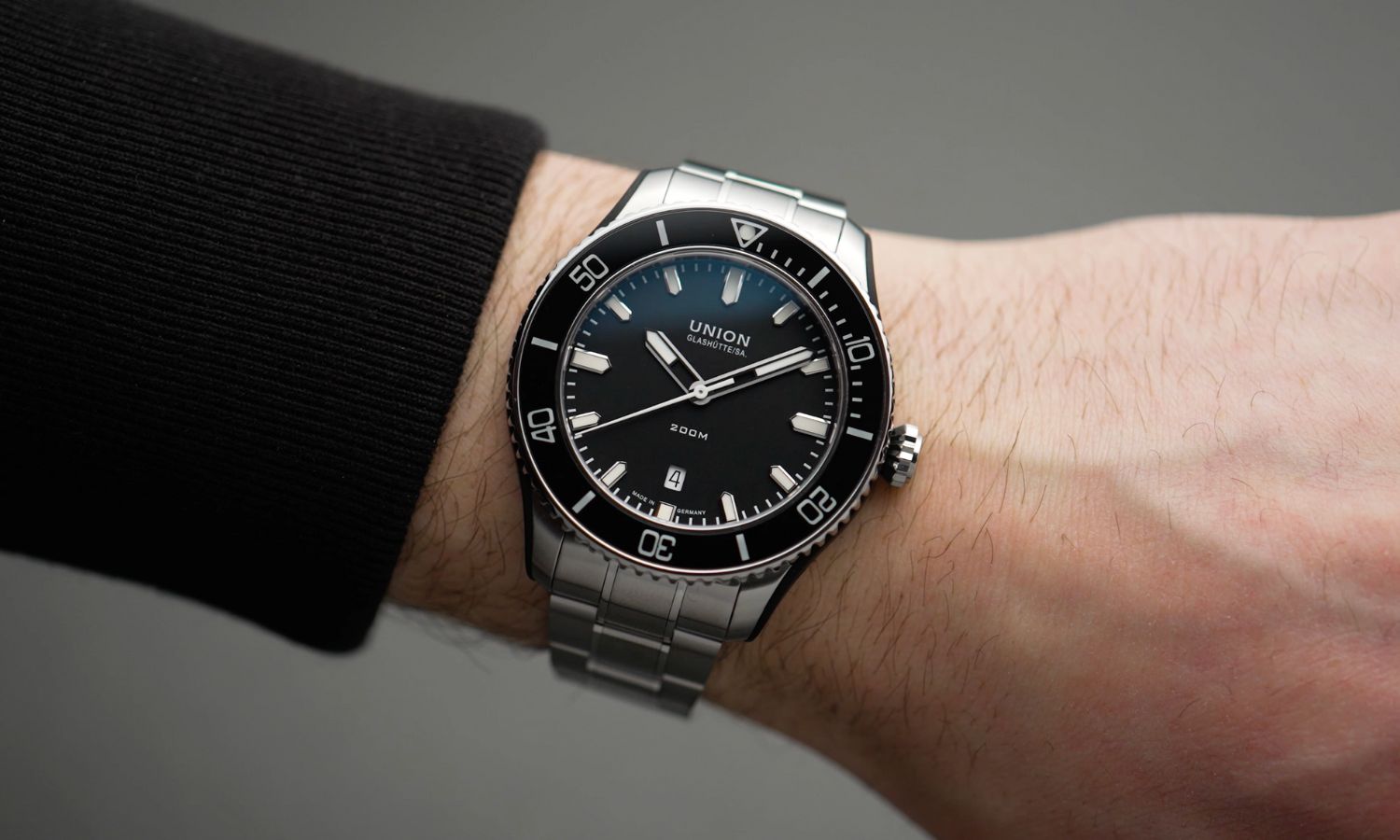 UNION Glashütte Belisar Datum Sport mit schwarzem Zifferblatt am Stahlband an einem Handgelenk mit schwarzem Ärmel getragen vor grauem Hintergrund