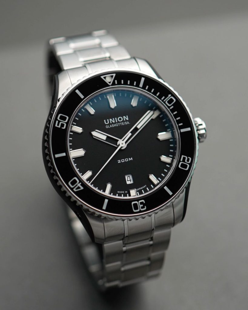 UNION Glashütte Belisar Datum Sport mit schwarzem Zifferblatt am Stahlband stehend vor grauem Hintergrund