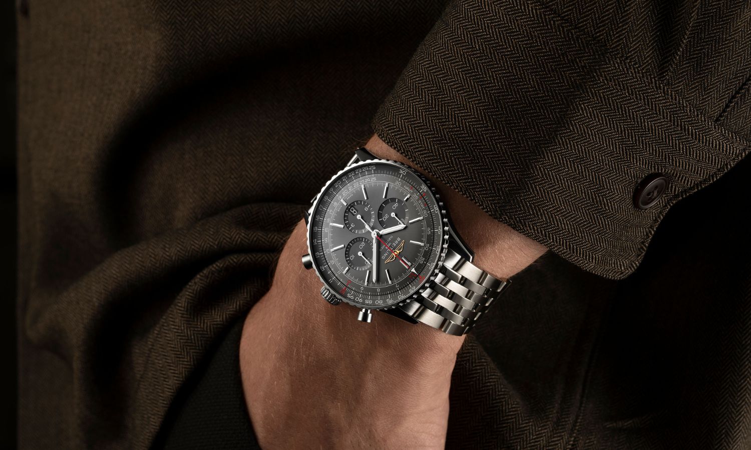 Breitling Navitimer Germany Edition mit anthrazitfarbenem Zifferblatt am Stahlarmband an einem Handgelenk mit braunem Ärmel getragen während die Hand in der Hosentasche ist