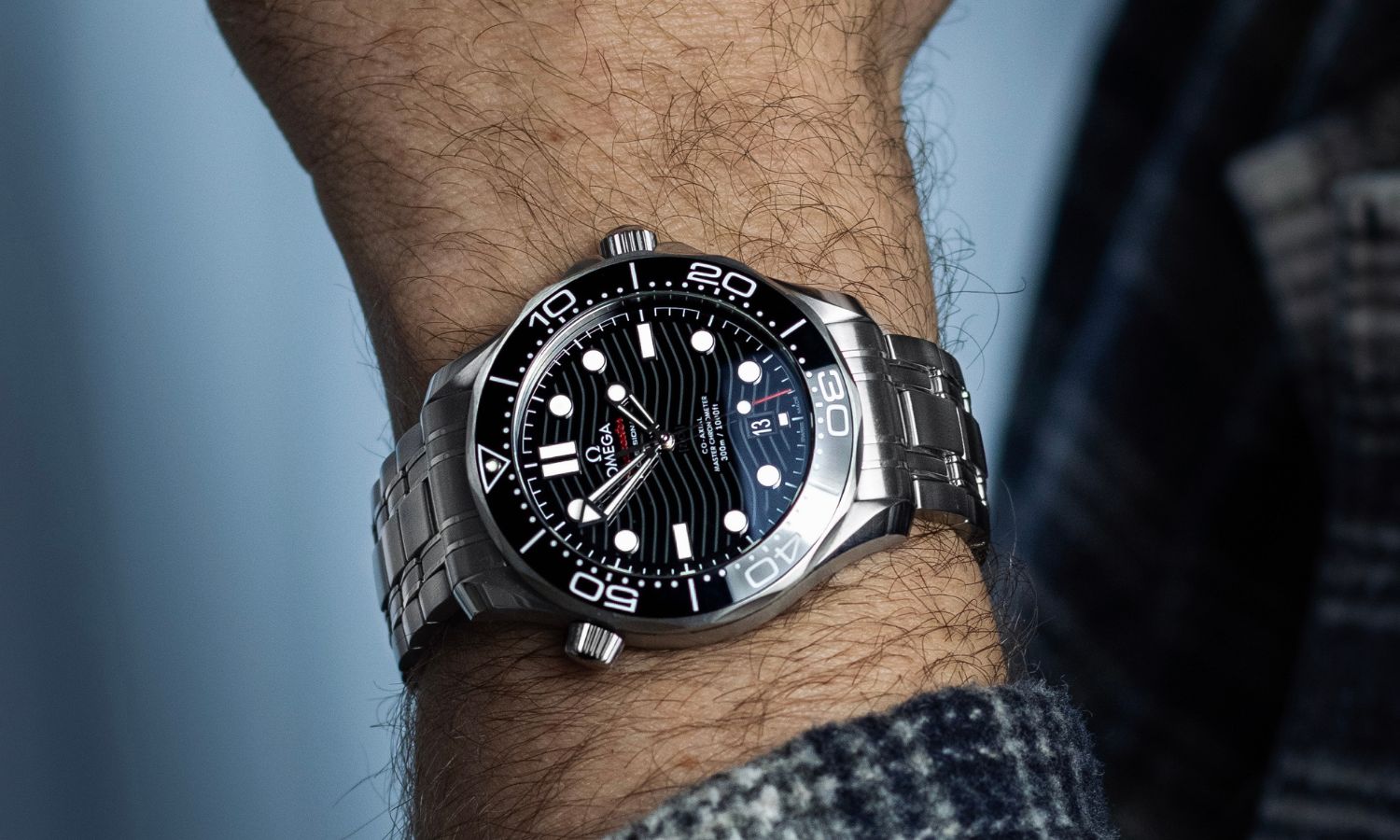 OMEGA Seamaster Diver 300M Referenz 210.30.42.20.01.001 mit schwarzem Zifferblatt am Edelstahlarmband an einem Handgelenk mit kariertem Ärmel getragen