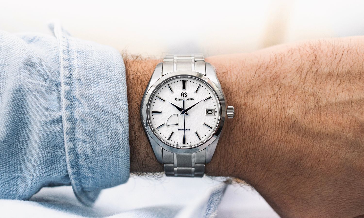 Grand Seiko Snowflake mit weißem Zifferblatt mit Titanarmband an einem Handgelenk getragen mit hellblauem Ärmel vor hellem Hintergrund