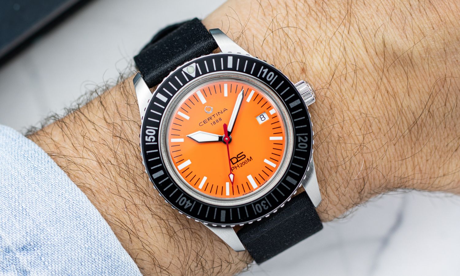 Certina DS PH200M Powermatic 80 mit orangenem Zifferblatt an einem Handgelenk getragen mit hellblauem Ärmel abgebildet vor hellem Hintergrund