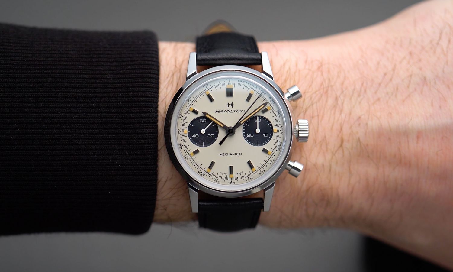 Der Hamilton Intra-Matic Handaufzug Chronograph mit silbernem Zifferblatt am schwarzen Lederarmband an einem Handgelenk mit schwarzem Ärmel getragen vor grauem Hintergrund
