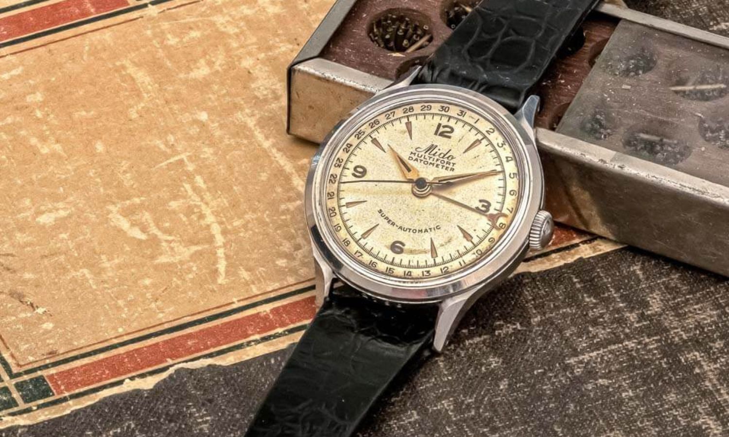 Mido Multifort Modell aus 1934 mit beigem Zifferblatt am schwarzen Lederarmband liegend abgebildet