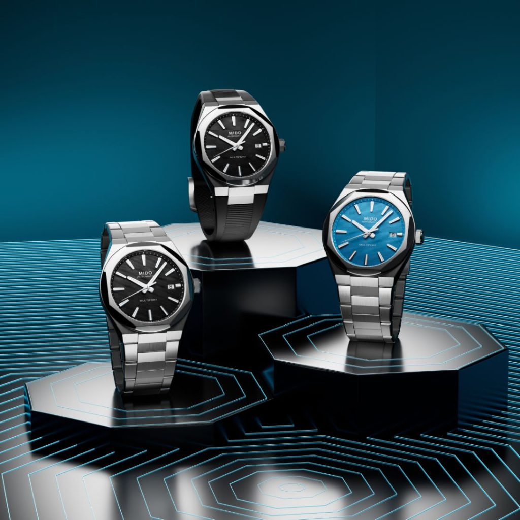 Mido Multifort 8 One Crown Referenz M055.507.11.051.00 mit schwarzem Zifferblatt am Edelstahlarmband, Mido Multifort 8 One Crown Referenz M055.507.17.051.00 mit schwarzem Zifferblatt am schwarzen Kautschukarmband und Mido Multifort 8 One Crown Referenz M055.507.11.041.00 mit blauem Zifferblatt am Edelstahlarmband stehend vor schwarz-blauem Hintergrund