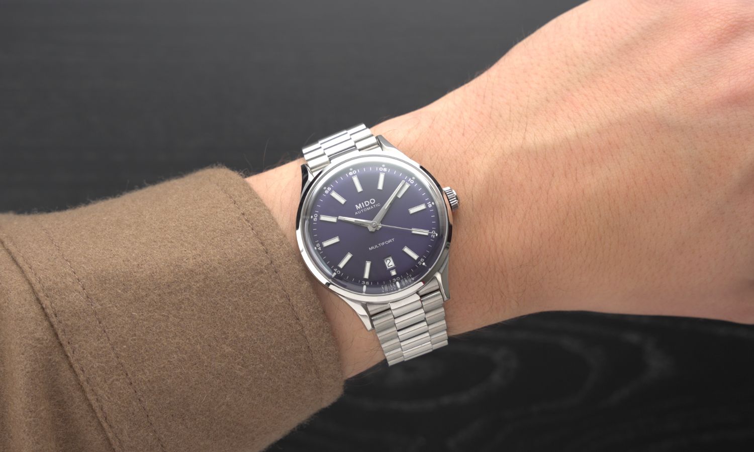Mido Multifort Patrimony Powerwind mit dunkelblauem Zifferblatt am Edelstahlarmband an einem Handgelenk mit hellbraunem Ärmel getragen vor schwarzem Hintergrund