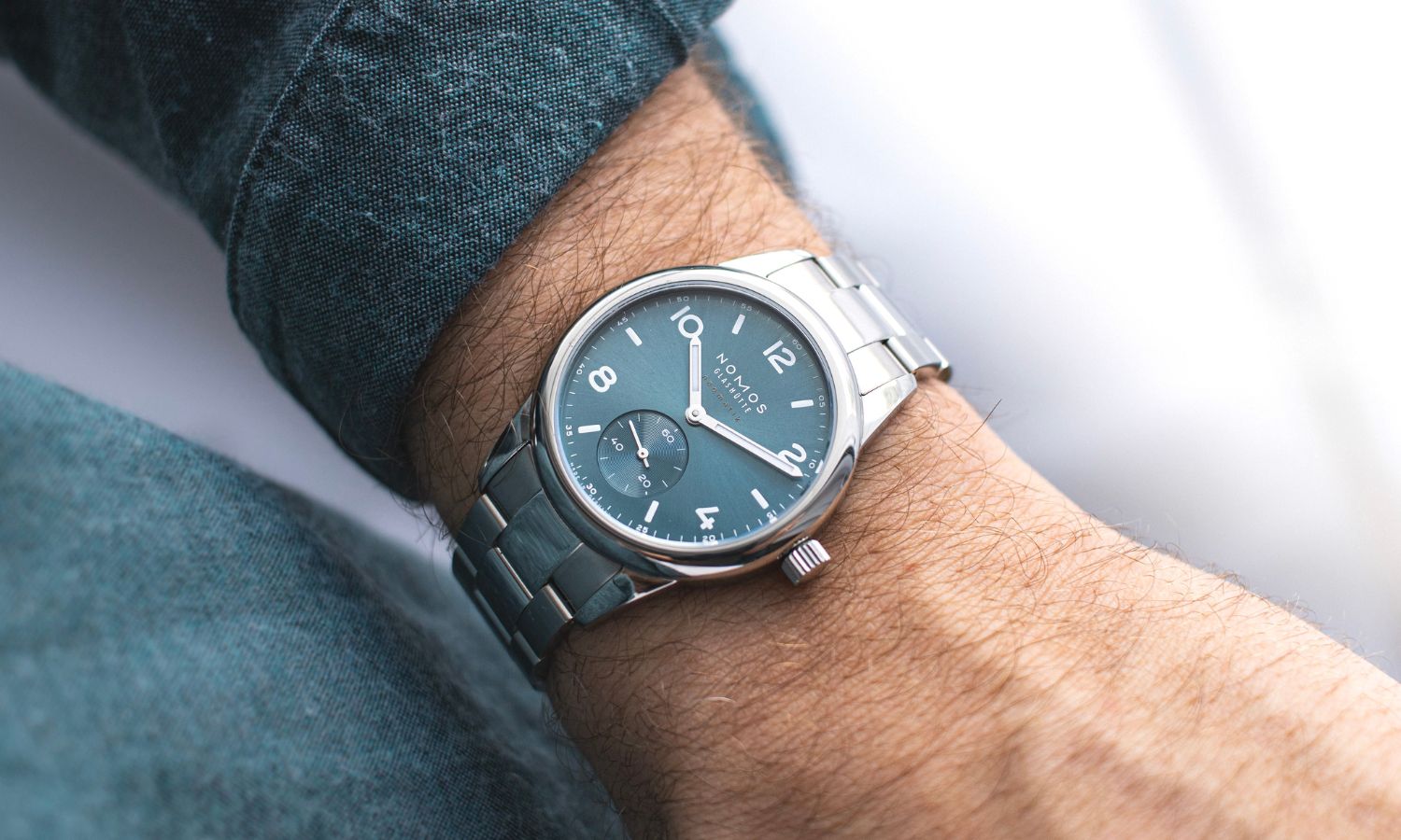 NOMOS Glashütte Club Sport neomatik mit petrolgrünem Zifferblatt am Edelstahlarmband an einem Handgelenk mit dunkelgrünem Ärmel getragen vor hellem Hintergrund abgebildet