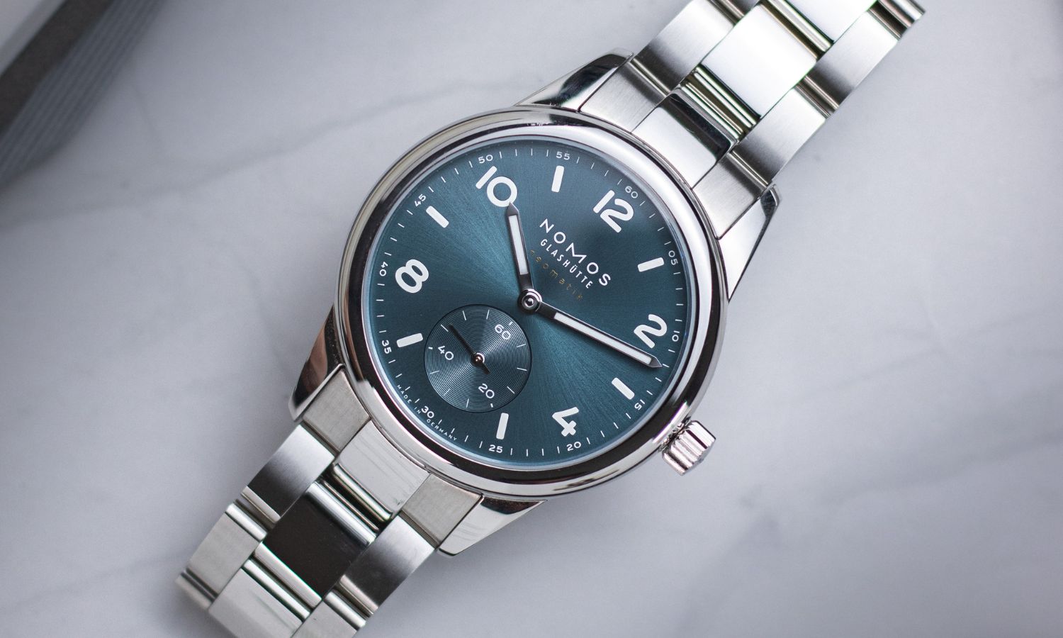 NOMOS Glashütte Club Sport neomatik mit petrolgrünem Zifferblatt am Edelstahlarmband seitlich liegend vor hellgrauem Hintergrund