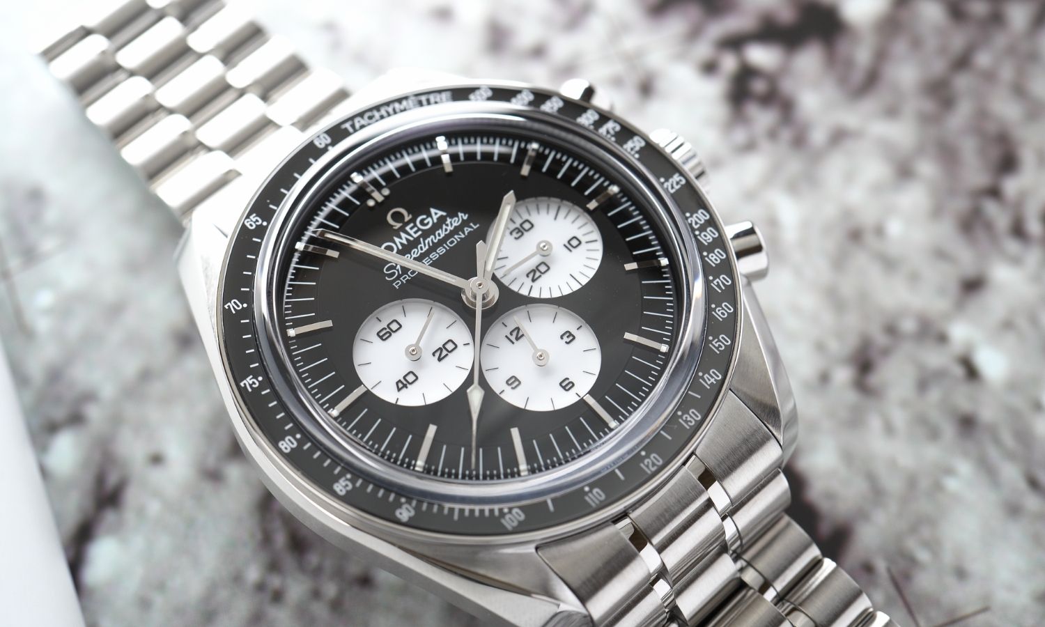 OMEGA Speedmaster Moonwatch Professional „Reverse Panda“ Referenz 310.30.42.50.01.004 mit schwarzem Zifferblatt am Edelstahlarmband abgebildet vor einem Mondoberflächen-Hintergrund