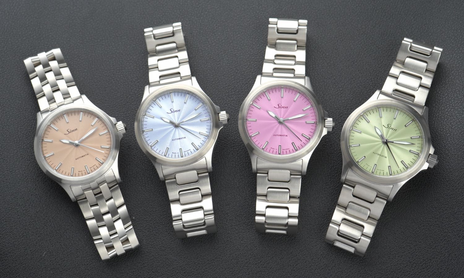 Sinn 556 H-Link Modelle mit sandfarbenem, eisblauem, pinken und grünem Zifferblatt nebeneinander liegend vor schwarzem Hintergrund