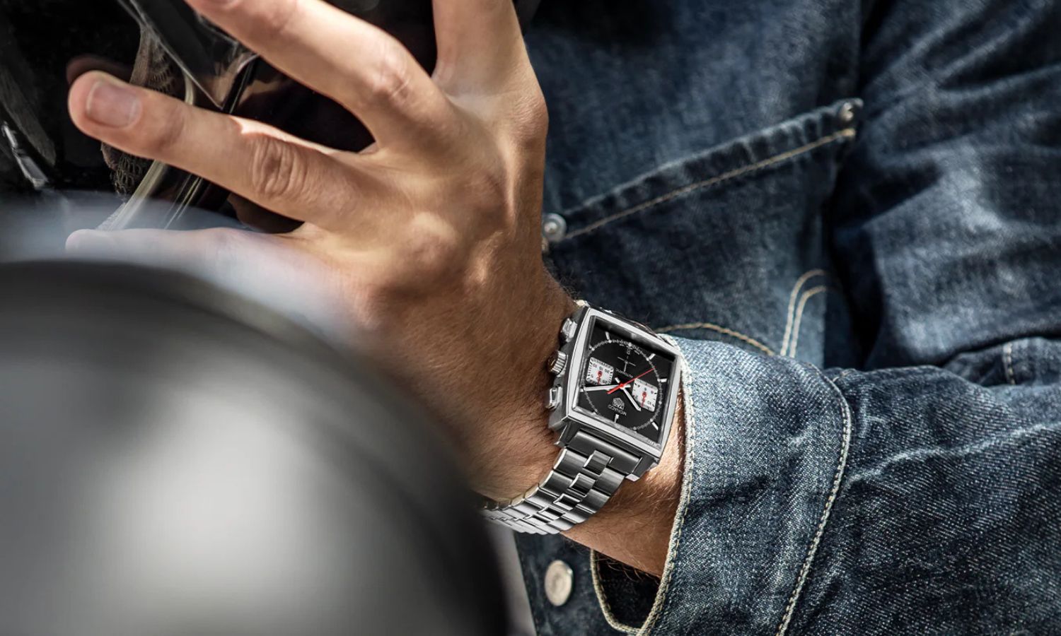 TAG HEUER Monaco Calibre Heuer 02 mit schwarzem Zifferblatt am Edelstahlarmband an einem Handgelenk eines Mannes mit Jeansjacke getragen