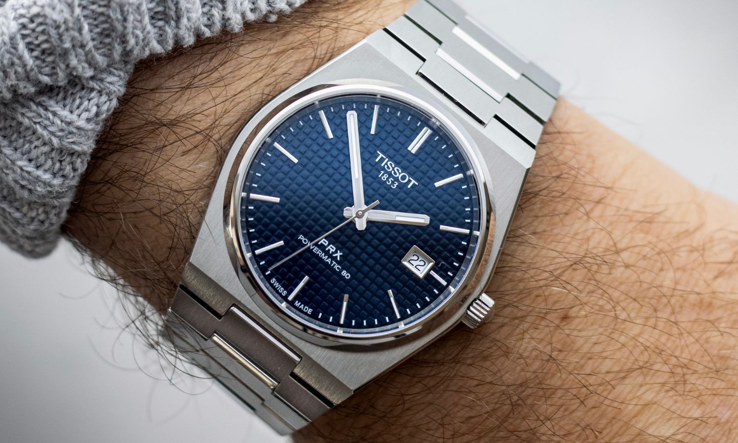 Tissot PRX Powermatic 80 mit dunkelblauem Zifferblatt am Edelstahlarmband an einem Handgelenk mit grauem Ärmel getragen vor hellgrauem Hintergrund
