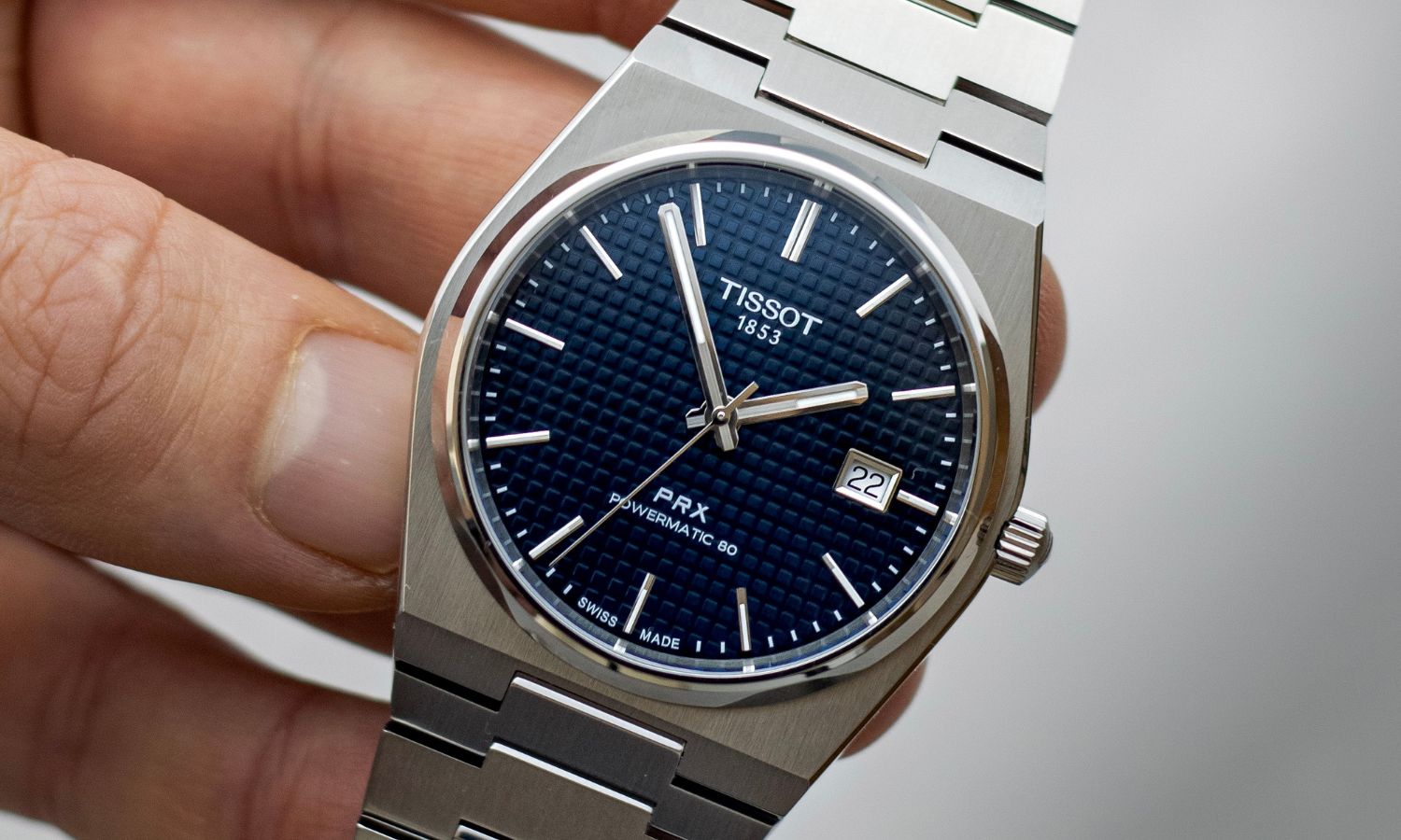 Nahaufnahme des dunkelblauen Zifferblatts der Tissot PRX Powermatic 80 am Edelstahlarmband in einer Hand gehalten vor hellgrauem Hintergrund