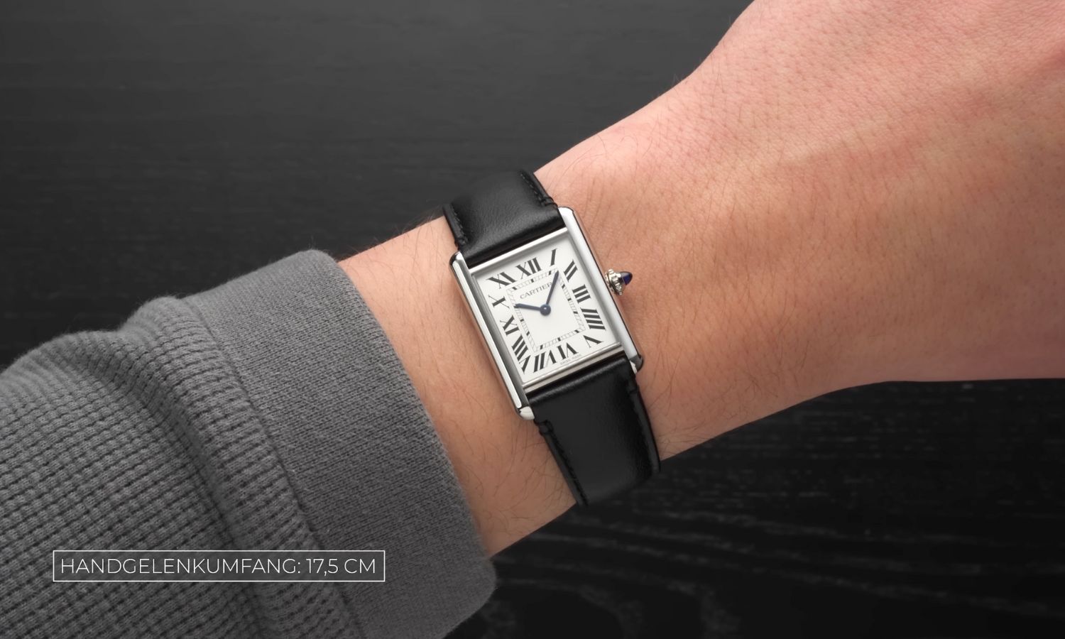 Cartier Tank Must SolarBeat mit silbernem Zifferblatt am schwarzen Lederarmband an einem Handgelenk mit grauem Ärmel getragen vor schwarzem Hintergrund