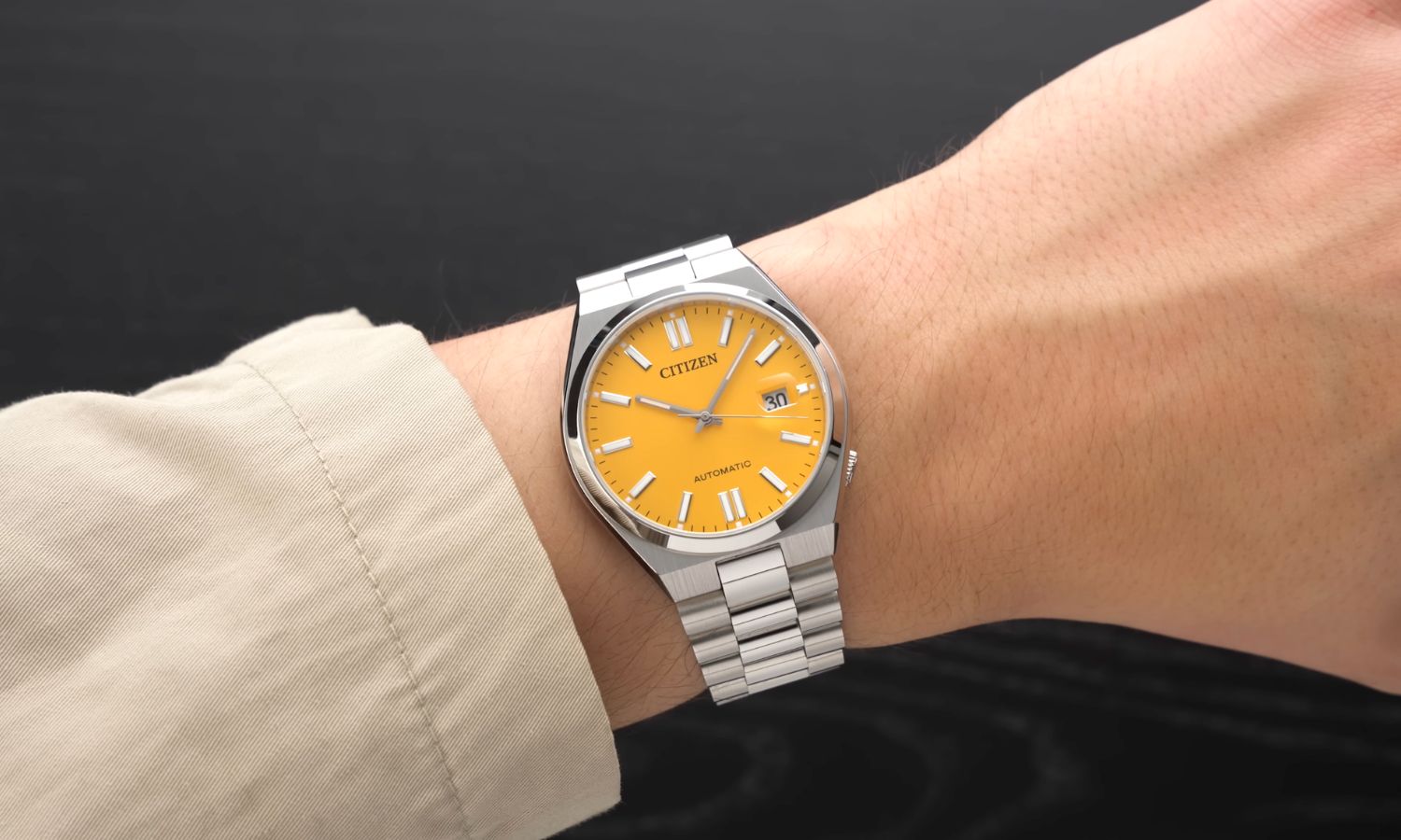 Citizen Tsuyosa mit gelbem Zifferblatt am Edelstahlarmband an einem Handgelenk mit beigem Ärmel getragen vor schwarzem Hintergrund