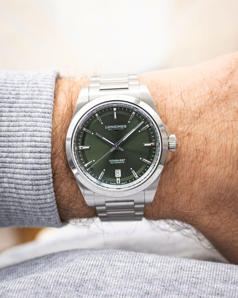Longines Conquest 38 mit grünem Zifferblatt am Edelstahlarmband an einem Handgelenk mit hellgrauem Ärmel getragen vor hellem Hintergrund abgebildet