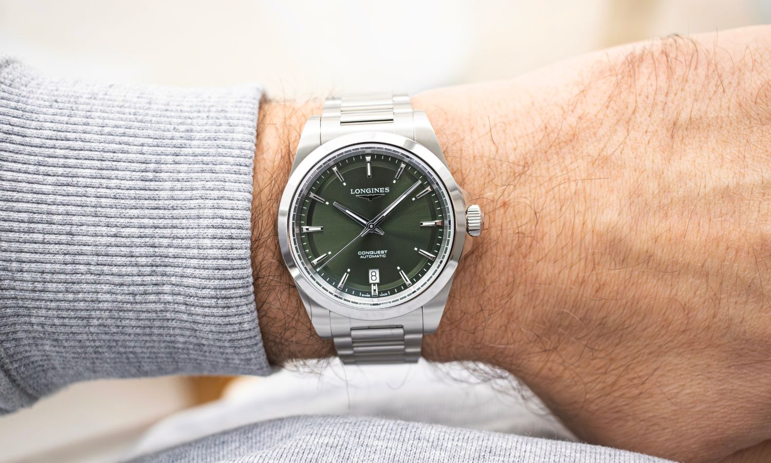 Longines Conquest 38 mit grünem Zifferblatt am Edelstahlarmband an einem Handgelenk mit hellgrauem Ärmel getragen vor hellem Hintergrund abgebildet