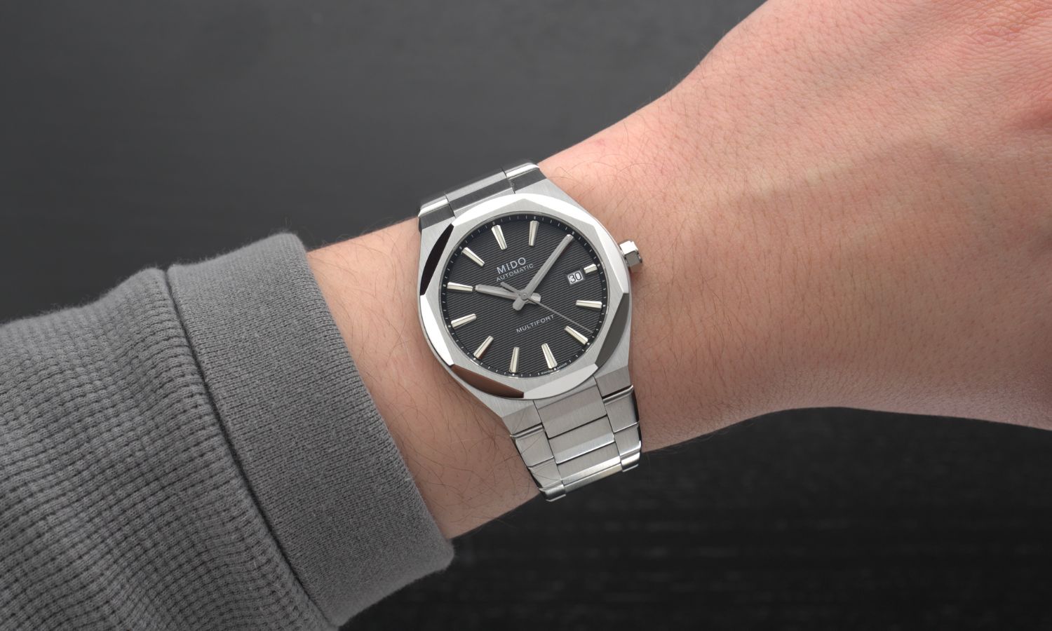Mido Multifort 8 One Crown Referenz M055.507.11.051.00 mit schwarzem Zifferblatt am Edelstahlband an einem Handgelenk mit grauem Ärmel getragen vor schwarzem Hintergrund