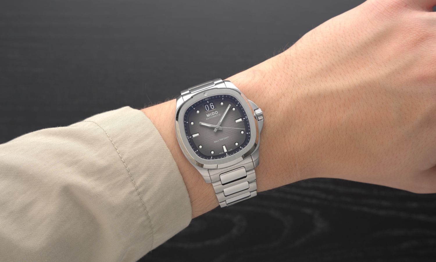 Mido Multifort TV Big Date Referenz M049.526.11.081.00 mit schwarzem Zifferblatt am Edelstahlband an einem Handgelenk mit beigem Ärmel getragen vor schwarzem Hintergrund