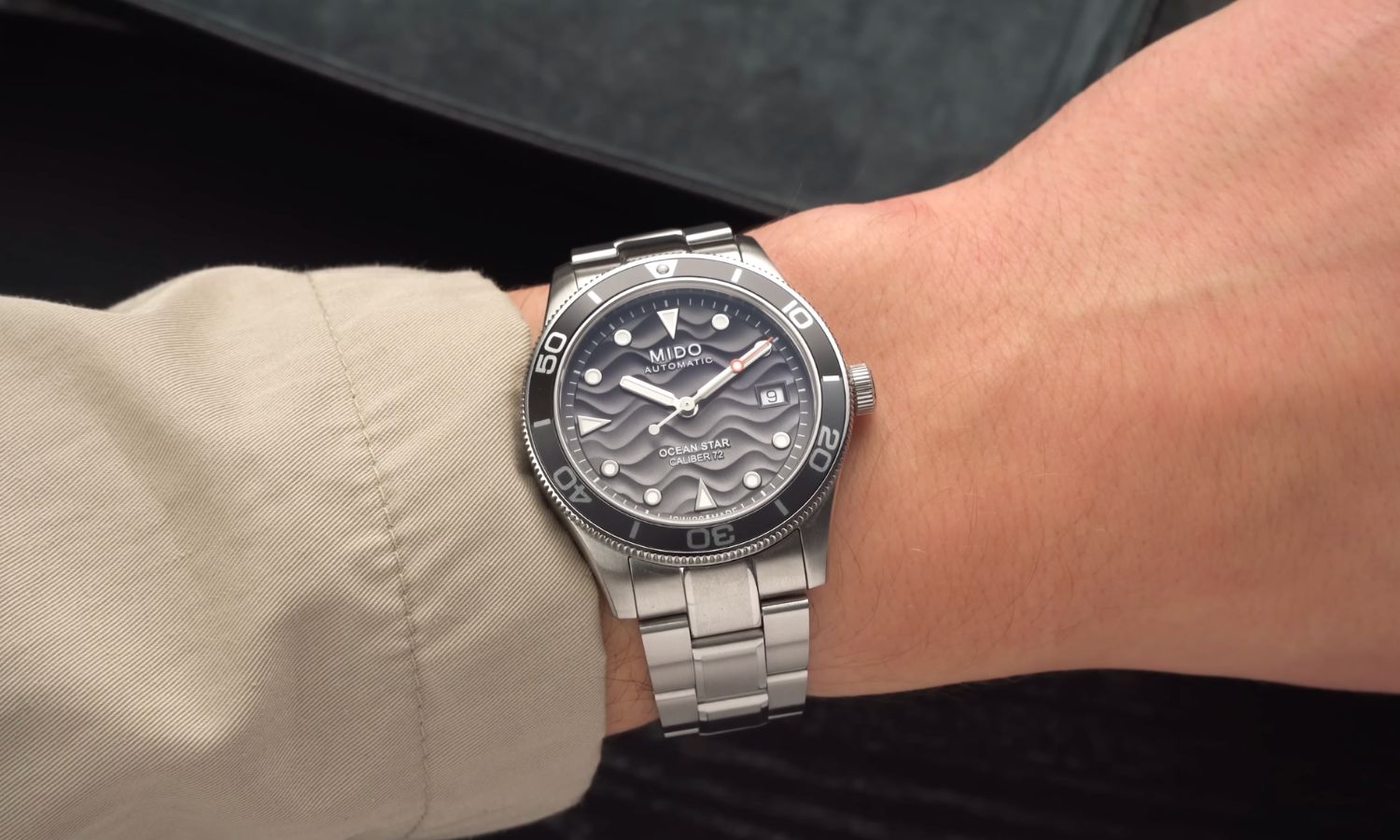 Mido Ocean Star 39 mit grauem Zifferblatt am Edelstahlarmband an einem Handgelenk mit beigem Ärmel getragen vor schwarz-grauem Hintergrund