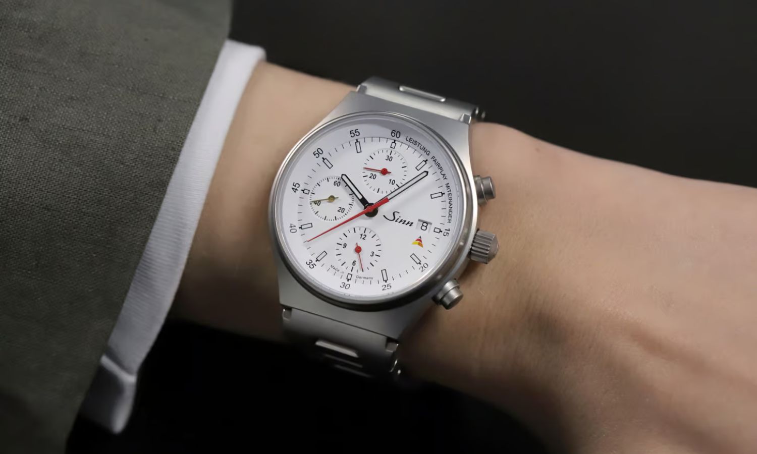 Sinn 144 St Sa Sporthilfe mit weißem Zifferblatt am Edelstahlarmband an einem Handgelenk mit grünem Ärmel getragen vor schwarzem Hintergrund