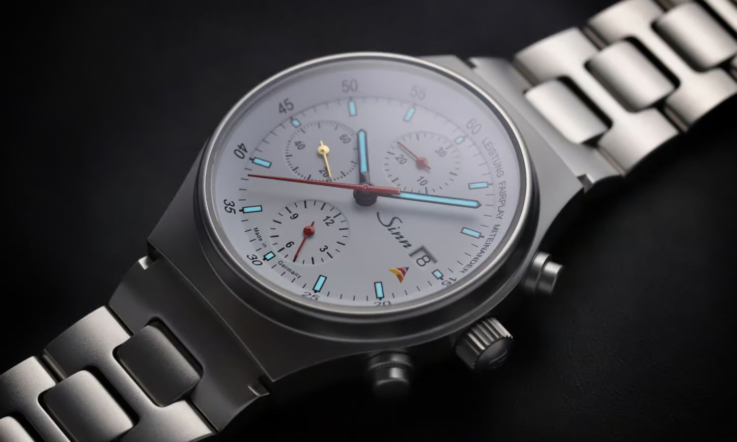 Sinn 144 St Sa Sporthilfe mit weißem Zifferblatt am Edelstahlarmband liegend auf schwarzem Untergrund