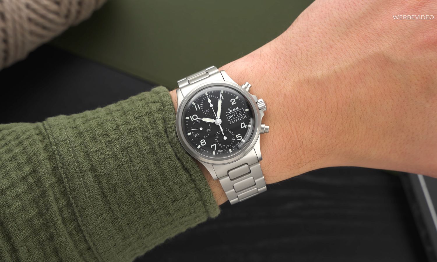 Sinn 356 Flieger mit schwarzem Zifferblatt am Edelstahlarmband an einem Handgelenk mit dunkelgrünem Ärmel getragen vor schwarzem Hintergrund abgebildet