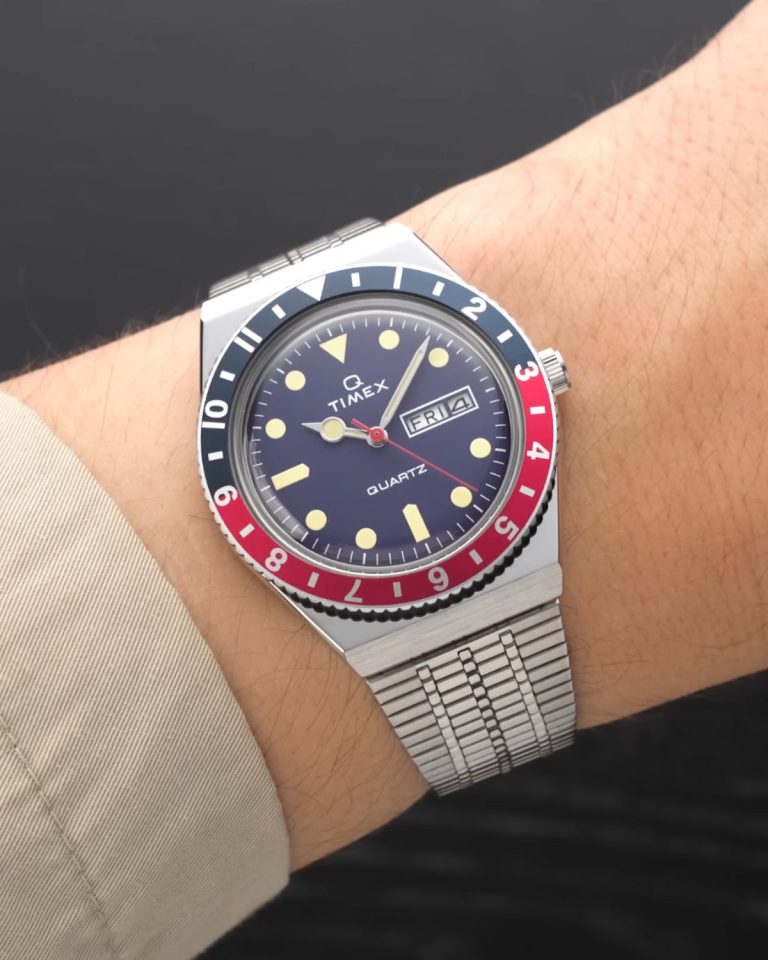 Timex Q Reissue mit dunkelblauem Zifferblatt am Edelstahlarmband an einem Handgelenk mit beigem Ärmel getragen vor schwarzem Hintergrund